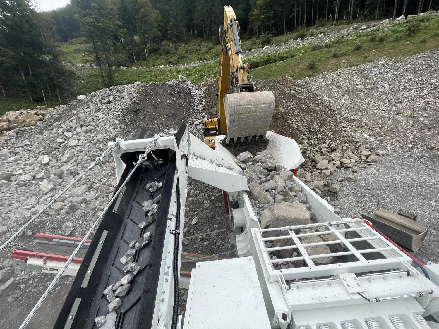 Förderband des mobilen Backenbrechers transportiert gebrochenes Material, Bagger im Hintergrund.