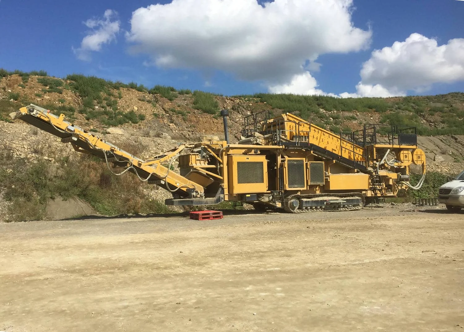 Mobile, gelbe raupenbasierte Brech- und Siebanlage im Einsatz auf einer staubigen Baustelle mit Vegetation im Hintergrund.