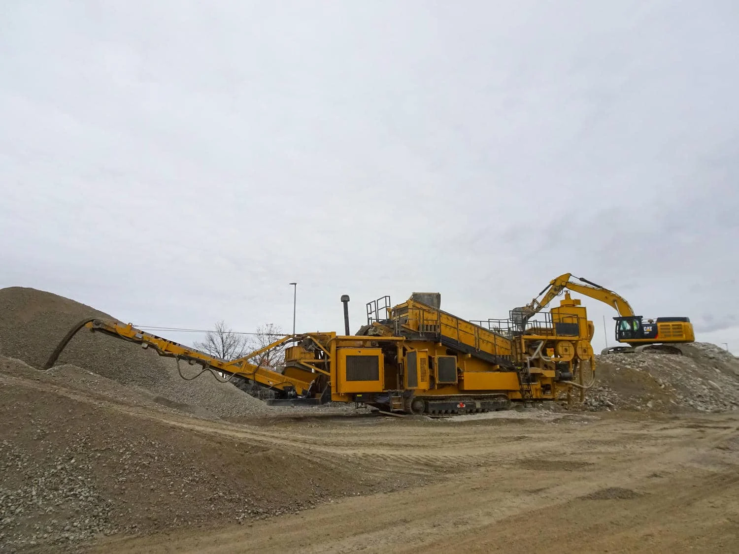Gelbe mobile Brech- und Siebanlage, die Material auf einem Kiesplatz verarbeitet, während ein Bagger die Maschine belädt.