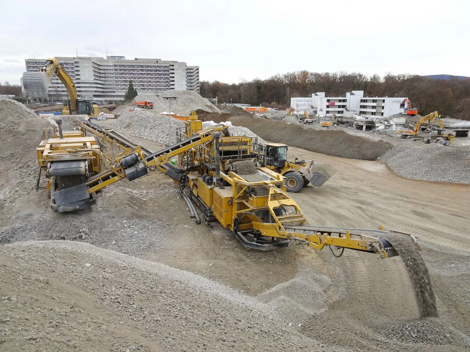 Großes Baustellengelände mit mehreren schweren Maschinen, darunter eine mobile gelbe Brech- und Siebanlage, ein Radlader und Bagger, die zwischen Kieshaufen nahe urbanen Gebäuden arbeiten.