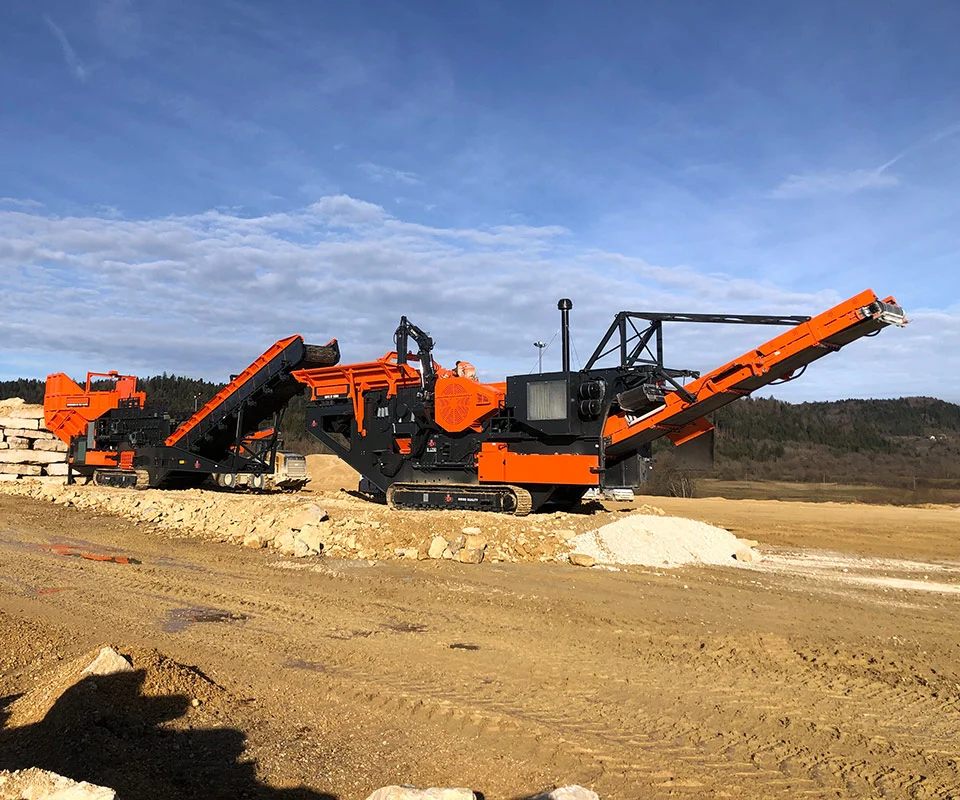 Mobile jaw crushing plant on tracks, processing blasted rock; discharge conveyors forming piles.